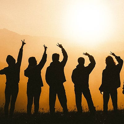 silhouette of people holding up peace sign in a field with the sung setting behind them.