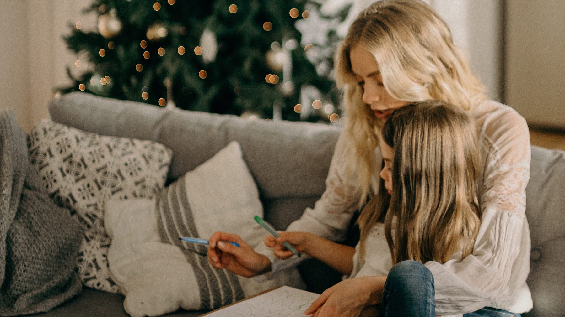 Mother and daughter drawing together on the couch with a Christmas tree in the background.